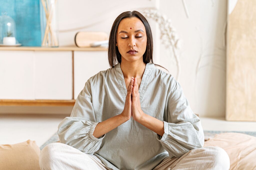 A woman is sitting cross-legged with her eyes closed, her hands clasped in front of her chest in prayer or as a greeting