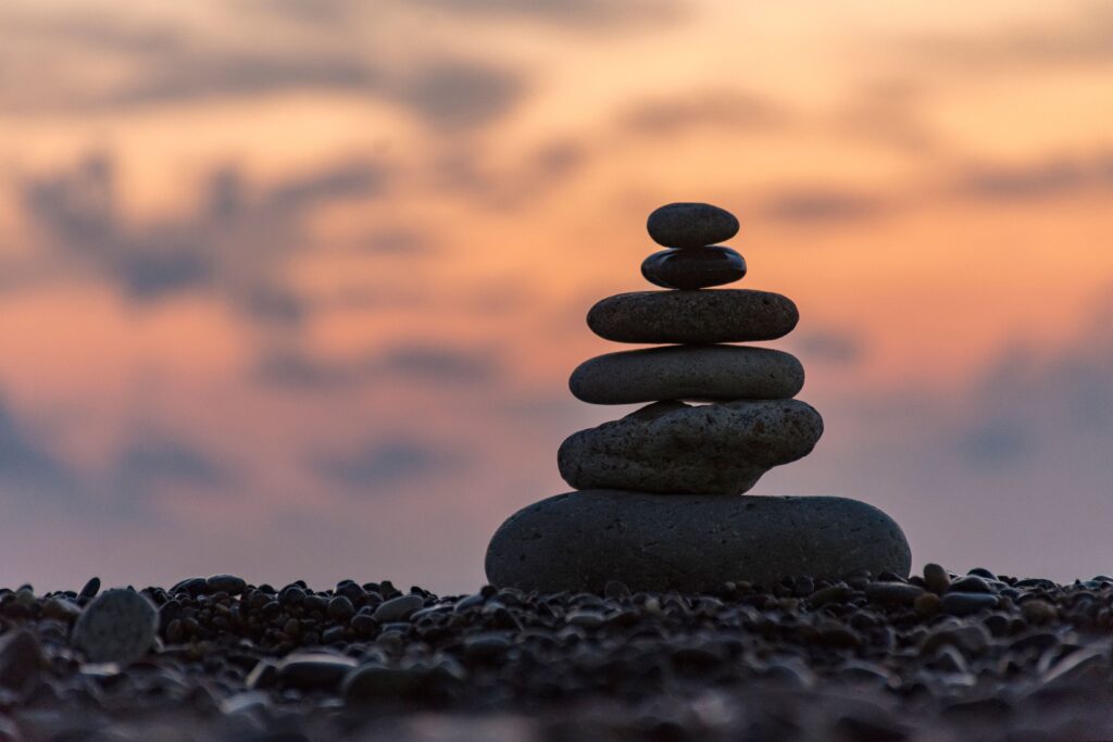 A pile of flat, dark stones—also known as a cairn—stands on a gravel beach against a colorful sunset sky.