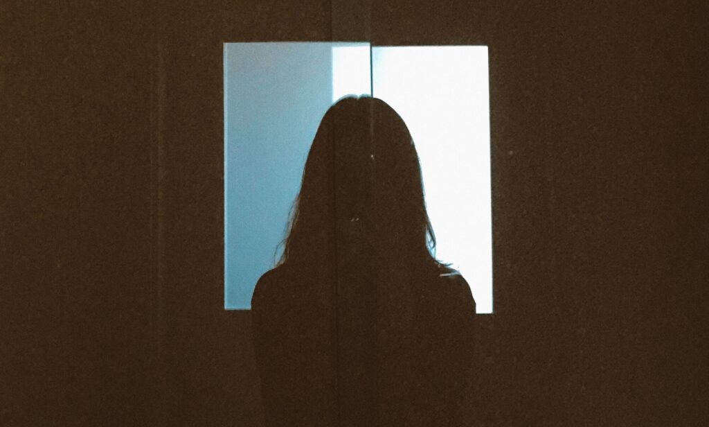 The silhouette of a person with long hair against a bright, square window