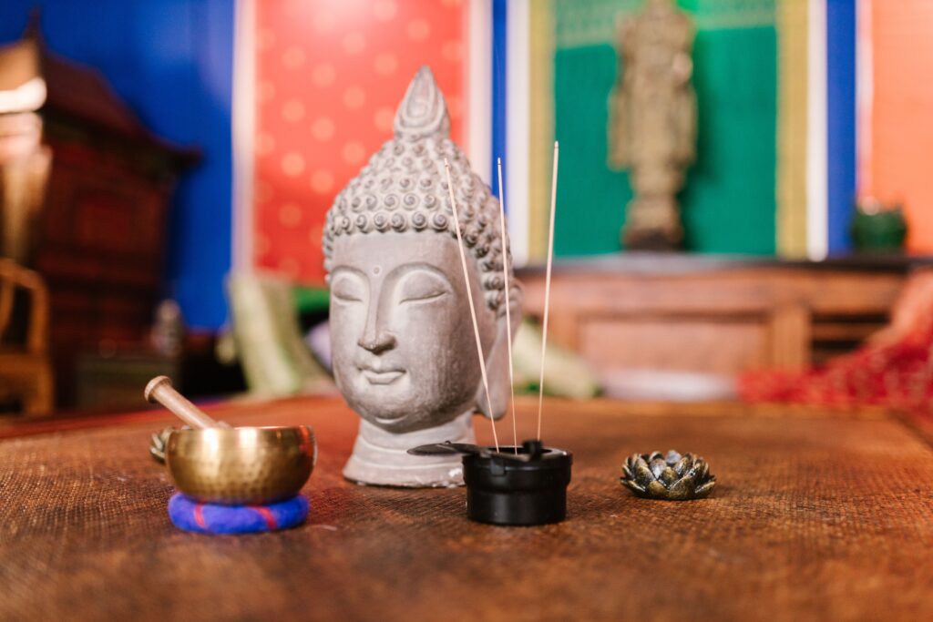 A bust of the Buddha, a singing bowl with a mallet, and incense sticks on a table against a colorful background.