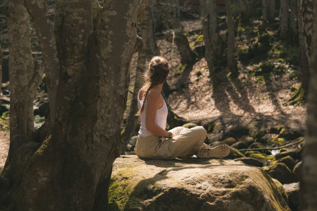 A woman is sitting with her back to the viewer on a large, moss-covered rock in the forest. She is wearing a white top and light brown pants. The scene is bright, and the ground is covered with leaves and stones.